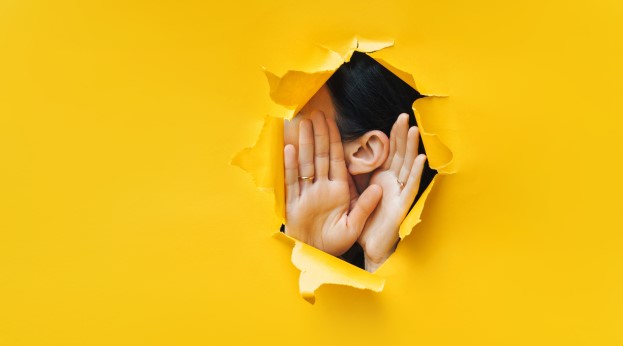 Listening through a hole in a yellow background with hands cupped around ear