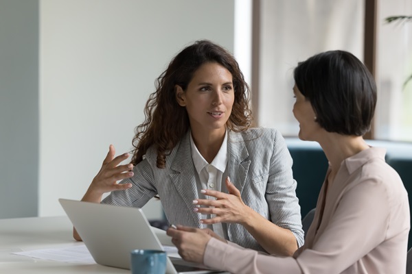 Two women talking at a desk with a laptop and a mug.