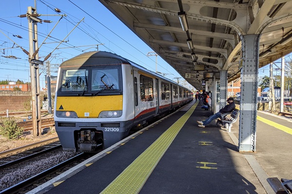 A train at Colchester railway station platform with a passenger waiting on a bench.