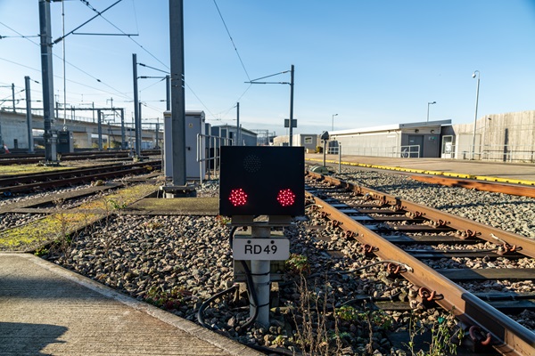 A flashing red LED warning light signal at a railway depot with tracks and overhead lines.
